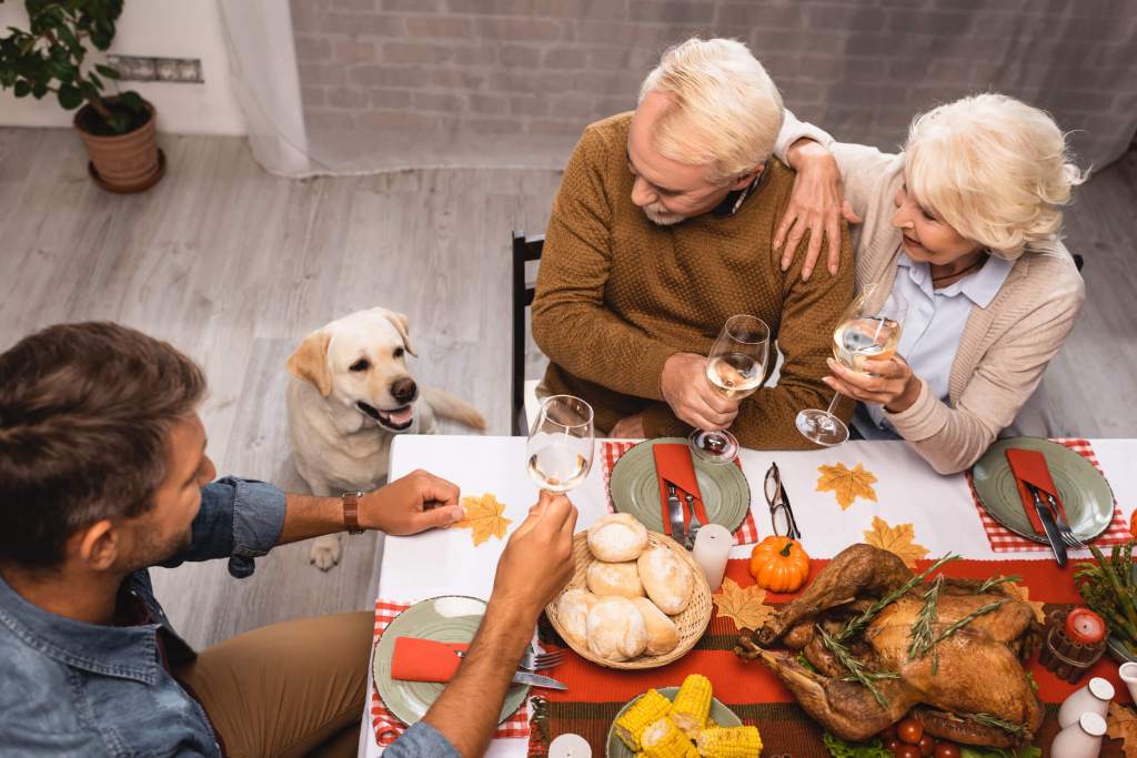 gettyimages-2177556455-1 Family eating Thanksgiving dinner with rolls and turkey on table. Senior couple to the right of a yellow Lab. Man to the left of dog.
